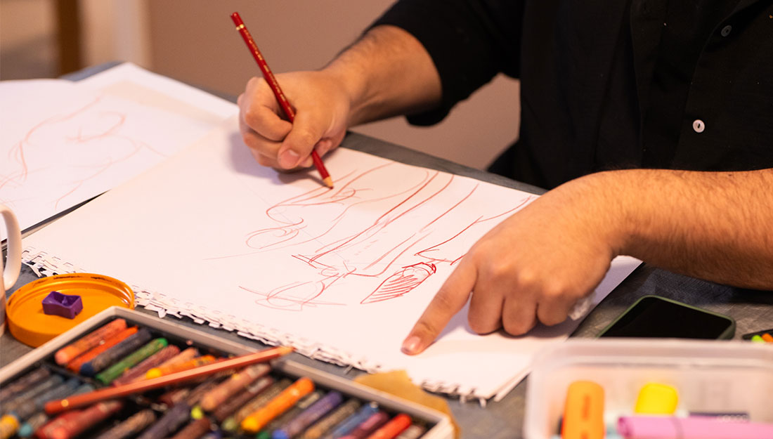 CraftingPathways_Abigail&Oly_PhotoHayleySalter_1100x625_3 A close-up of Abigail drawing three figures in red pencil. They are abstracted.