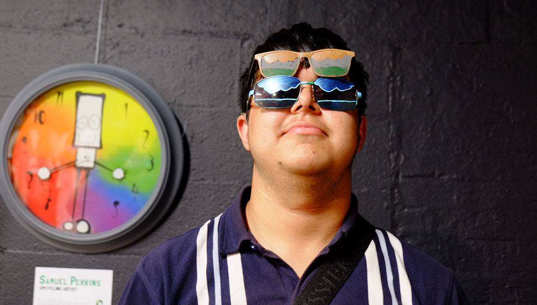 Braidwood_ExhibGlasses2 A young man wears two pairs of sunglasses. One decorated with dark colours and one with bright.